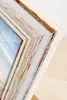A close-up view of a distressed white and brown wooden picture frame with a blue and white image visible inside. Produktbild-Vorschau