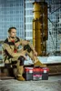A man in work clothes sits on a metal beam with a toolbox in front of him, with a construction site in the background. Produktbild-Vorschau