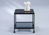 A black metal side table with a glass shelf and wheels, decorated with two white decorative flowers in vases on top. Produktbild-Vorschau