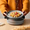 A person in an orange sweater is holding a plate of food over a stack of plates. Produktbild-Vorschau