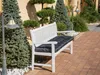 A white garden bench with a black cushion sits on a brick patio, surrounded by evergreen trees and decorative shrubs. Produktbild-Vorschau
