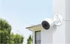 A white security camera is mounted on a white wall, with a house and trees in the background. Voir les images du produit
