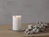 A white LED candle sits on a glass coaster on a wooden surface, next to a branch with cotton bolls. The background is a plain, textured gray wall. Podgląd zdjęcia produktu