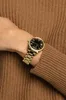 A person's wrist wearing a gold watch with a black face, against a brown knitted sweater. Náhled obrázku produktu