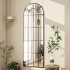 A tall, arched mirror with a black grid frame is placed against a textured beige wall. Sunlight casts striped shadows on the wall. Plants are placed on either side of the mirror, and a pendant light hangs from the ceiling. Produktbild-Vorschau