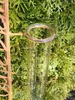 A close-up of a glass rain gauge with measurement markings, attached to a decorative metal stand, set against a backdrop of green foliage. Náhled obrázku produktu