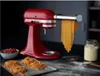 A red KitchenAid stand mixer with a pasta attachment is making fresh pasta. There is a bowl of chopped tomatoes and a jar of flour in the background, with freshly made pasta on a tray in the foreground. Produktbild-Vorschau