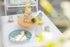 A table set for a meal outdoors with plates, cutlery, glasses, and a pitcher of water with lemon. Anteprima dell’immagine prodotto