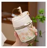 A hand holding a glass water bottle with a floral sleeve and a straw lid. Podgląd zdjęcia produktu