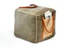 A cube-shaped pouf with leather handles and a front pocket containing a newspaper and glasses. Náhľad obrázku produktu
