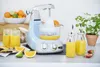 A light blue stand mixer with a juicer attachment is shown on a kitchen counter, surrounded by fresh oranges, limes, strawberries, and glasses of orange juice. Anteprima dell’immagine prodotto