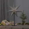 A large, grey, star-shaped lamp is displayed on a wooden surface, surrounded by wrapped gifts and small fairy lights. A miniature Christmas tree in a glass jar is also present, creating a festive, indoor holiday scene. Podgląd zdjęcia produktu