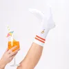A person's legs wearing white socks with orange stripes and a drink in a glass. Produktbild-Vorschau