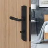 A black door handle set on a wooden door, with a bedroom interior visible in the background. Produktbild-Vorschau