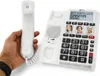 A hand holding the handset of a white corded telephone with a large keypad and a digital display. Náhľad obrázku produktu