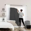 A woman is hanging a dark gray blackout curtain in a bedroom window, with a suitcase and bag next to the window. Produktbild-Vorschau