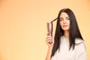 A woman holds a curling iron with a strand of her hair inside it against a plain orange background. Náhled obrázku produktu