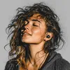 A woman with curly hair is wearing earbuds and enjoying the sun. Podgląd zdjęcia produktu