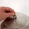 A hand is shown adjusting a small brass valve on the lid of a stainless steel appliance, likely a pressure cooker or press. Produktbild-Vorschau