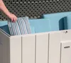 A hand is placing a striped cushion into a beige storage box filled with blue cushions. Podgląd zdjęcia produktu