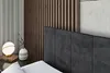A close-up view of a bed with a dark upholstered headboard, a white pillow, and a bedside table with a lamp and a vase. Produktbild-Vorschau