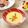 A close-up shot of a red and white patterned soup bowl filled with yellow soup, garnished with orange roe and green herbs. In the background, a plate of sliced meat and a bowl of fruit salad are visible. Produktbild-Vorschau