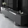 A modern TV cabinet with a sleek design, featuring a dark gray finish and silver accents. It is placed in a dimly lit room, with a large black television screen visible in the background. Voir les images du produit