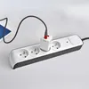 A white and black power strip with four outlets is shown on a light gray surface. A white power adapter is plugged into one of the outlets, with a black charging cable connected to it and extending towards a blue object on the left. Náhľad obrázku produktu