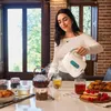 A woman is pouring hot water from an electric kettle into a glass cup, preparing tea. There are various tea accessories and pastries on the table. Produktbild-Vorschau