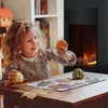 A young girl is sitting at a table, holding small pumpkins and arranging leaves on a piece of paper. A fireplace is visible in the background. Produktbild-Vorschau