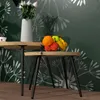 A close-up view of two wooden side tables with black metal legs, one holding a vase of roses and a small cup, the other holding a bowl of fruit. The background is a dark green wall with a white dandelion-like pattern. Produktbild-Vorschau