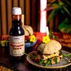 A bottle of Worcester sauce next to a burger, with decorative elements. Produktbild-Vorschau