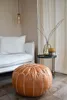 A Moroccan leather pouf sits on a wooden floor next to a white sofa, with a small side table holding a vase and candle holder in the background. Voir les images du produit