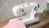 A white sewing machine is shown on a wooden table, with a piece of pink fabric being sewn. Produktbild-Vorschau