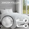 A white comforter is shown on a bed with two pillows, with a magnifying glass highlighting the filling. A window with trees is visible in the background. Produktbild-Vorschau