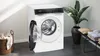 A white Siemens washing machine with its door open, placed in a modern laundry room setting with a basket of clothes and a potted plant. Produktbild-Vorschau