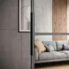 A modern living room interior with a sofa and wooden wall paneling, viewed through a glass door with a black handle. Produktbild-Vorschau