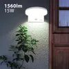A white LED wall light with a motion sensor is mounted on a textured wall next to a wooden door. A green plant is in the foreground, illuminated by the light. Voir les images du produit