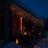 Three people sitting at a table outside a wooden structure at night, illuminated by colorful icicle lights and a candle. Produktbild-Vorschau