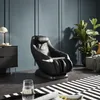 A black massage chair is placed in a modern living room setting, next to a dresser and a large potted plant. Produktbild-Vorschau