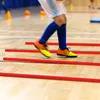 A person is shown performing agility drills on a wooden floor with red agility poles. Náhled obrázku produktu