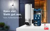 A Bosch Eyes Außenkamera II is shown on a white surface next to a smartphone displaying the camera's app interface. The background shows a house exterior with a light and a door. Anteprima dell’immagine prodotto