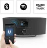 A person is holding a smartphone in front of a black Hama DAB radio. The radio has a screen displaying music information and several buttons. Above the radio are three square icons representing Bluetooth, Spotify, and Amazon Music. Produktbild-Vorschau