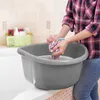 A person is washing a colorful cloth in a gray basin filled with soapy water, placed on the edge of a bathtub. Produktbild-Vorschau