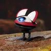 A small, red and white robot with blue glowing eyes stands on a rocky surface with a blurred brown background. Anteprima dell’immagine prodotto