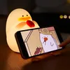 A glowing duck-shaped night light is holding a smartphone that displays a cartoon of a duck wearing a straw hat. Produktbild-Vorschau
