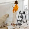 A woman is painting a wall with a roller, while a large dog sits nearby. A black stepladder is positioned to the right of the woman. Voir les images du produit