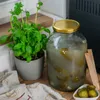 A large glass jar filled with pickles and brine, next to a potted basil plant and a small bowl of pickles. Podgląd zdjęcia produktu