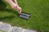 A person's hands are shown using a tool to adjust a black plastic box with multiple compartments, which is placed on a green lawn next to a stone pathway. Produktbild-Vorschau