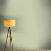 A floor lamp with a wooden tripod stand is placed on a wooden floor against a plain green wall. Produktbild-Vorschau